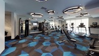This fitness center offers a comprehensive, well-appointed workout facility designed with both functionality and aesthetics in mind. The space is distinguished by its striking geometric-patterned carpet in shades of blue, gray, and charcoal, paired with a contemporary gray color palette on the walls and ceiling. Overhead, an array of bold circular ring pendant lights provides ample, stylish illumination throughout the floor.

The main workout area is generously sized, housing a full lineup of cardio equipment including multiple treadmills, elliptical trainers, and stationary bikes, all equipped with individual digital displays. Large windows along one wall are fitted with roller shades, allowing natural light to filter into the space. Full-length mirrors on one side of the room visually expand the area and support strength training activities.

A dedicated TRX Training Zone occupies a distinct section of the facility, outfitted with suspension trainers, kettlebells, medicine balls, and a rowing machine, set against a textured accent wall for an energetic atmosphere. A separate free weights area features a dumbbell rack and adjustable bench, accompanied by a wall-mounted flat-screen television. Lockers and a water station are conveniently accessible, rounding out the practical amenities of this thoughtfully designed fitness center.
