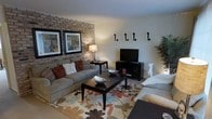 This single-floor space centers around a well-appointed living area that immediately draws attention with its textured exposed brick accent wall, which serves as a dramatic backdrop for the room's stylish furnishings. The living area is anchored by a plush, neutral-toned sectional sofa adorned with an assortment of decorative throw pillows in warm earth tones, complementing the overall color palette of the space. A dark-finished coffee table sits atop a large, eye-catching area rug featuring a bold floral pattern in shades of brown, rust, and cream, adding visual interest to the room. The entertainment setup includes a flat-screen television positioned on a matching dark media console, keeping the space functional and organized. Ambient lighting is provided by a classic table lamp, casting a warm glow that enhances the inviting atmosphere. Decorative wall art and ornamental accents, including sculptural wall hooks and tasteful framed prints, add personality and a curated feel to the walls. A lush indoor plant in the corner introduces a touch of greenery, while a decorative figurine adds a whimsical detail. The overall ambiance is warm, lived-in, and thoughtfully styled, making this space feel both comfortable and visually engaging.