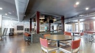 This single-floor space presents a contemporary dining and lounge environment defined by a striking design aesthetic. The dark, dramatic ceiling creates an intimate atmosphere, while exposed structural columns clad in rich brick and deep wood tones add an industrial-chic character throughout the space. Pendant lighting with square drum shades hangs above key areas, casting a warm, focused glow across the room. A central island or bar counter finished in stacked stone serves as the focal point, offering bar-height seating with bold orange upholstered stools that inject a vivid pop of color against the neutral palette. Round dining tables paired with matching orange and tan cushioned chairs are arranged to encourage social interaction and comfortable dining. Wall-mounted flat-screen televisions are positioned for easy viewing from multiple seating areas. Decorative wall art and a large-scale mural or textured wall treatment add visual interest and personality to the space. Light wood-toned flooring grounds the room and contrasts beautifully with the darker overhead elements. The open layout ensures easy flow between seating zones, creating a welcoming, versatile environment suited for dining, socializing, or community events.