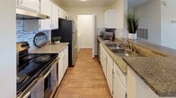 This single-story space showcases a well-appointed galley-style kitchen designed for efficient use of space. The layout features cabinetry lining both sides of the kitchen, finished in crisp white with ample storage above and below the counters. The countertops offer a granite-look surface in warm earth tones, providing generous prep space along both walls.

On one side, a black electric range and a stainless steel refrigerator anchor the cooking area, complemented by an over-the-range microwave. The opposite wall features a double stainless steel sink with a standard faucet, ideal for food preparation and cleanup. A decorative stone-look tile backsplash runs along one wall, adding a textural accent that enhances the kitchen's visual interest.

The warm wood-look flooring runs the length of the kitchen, contributing to the inviting atmosphere. At the far end of the kitchen, a doorway leads to an adjacent area, suggesting a practical flow throughout the space. The kitchen is well-lit with overhead lighting, ensuring a bright and functional environment.