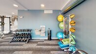 Located at 2850 Uptown Way S in Fargo, North Dakota, this well-appointed fitness center delivers a comprehensive workout environment designed to meet a wide range of exercise needs. The space is flooded with natural light through expansive floor-to-ceiling windows that line the cardio area, offering open views of the surrounding Uptown Fargo landscape and creating an energizing atmosphere for exercise.

The cardio section is generously equipped with a row of treadmills, elliptical machines, and stationary bikes, each positioned to take advantage of the outdoor views. Wall-mounted flat-screen televisions are stationed above the cardio equipment, providing entertainment during workouts. Recessed lighting throughout the space ensures consistent illumination alongside the abundant natural light.

A dedicated strength training area features a full power rack loaded with Olympic barbells and weight plates, catering to serious lifters. The free weights section includes a tiered dumbbell rack with a varied selection of weights, ideal for resistance training. A glass-enclosed room provides additional workout space, visible from the main floor.

The functional training corner is stocked with medicine balls, resistance bands, stability balls, exercise mats, and a dual water fountain station, making it a versatile area for stretching and cross-training. Patterned carpet tiles in neutral tones cover the flooring throughout, tying together the clean, modern aesthetic of this well-rounded fitness facility.