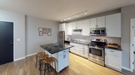 This 2-bedroom, 2-bathroom model residence delivers a thoughtfully designed single-floor layout with contemporary finishes and a cohesive, modern aesthetic. The open-concept kitchen is a standout feature, showcasing white shaker cabinetry, dark granite countertops, a mosaic tile backsplash, stainless steel appliances, and track lighting overhead. A movable kitchen island with bar seating adds both function and flexibility. Rich hardwood flooring flows through the kitchen and living areas, creating a warm, connected feel. The living room is spacious and bright, benefiting from large sliding glass doors that open to a private balcony with outdoor views, filling the space with natural light. Both bedrooms are carpeted for comfort, featuring neutral gray walls and windows that invite additional natural light. The two bathrooms are finished with wood-look flooring, white vanities with dark hardware, large mirrors, and clean subway tile detailing in the shower areas — offering a crisp, modern look. The overall color palette of soft grays, whites, and warm wood tones creates a calm and inviting ambiance throughout. The layout is efficient and well-proportioned, making excellent use of the available square footage across every room.