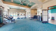 The fitness room at Canopy Apartment Villas in Pelham, Alabama, is a thoughtfully designed, single-level facility offering a comprehensive workout environment. The main exercise area features elevated tray ceilings accented in teal, recessed lighting, and ceiling fans that contribute to an open, airy atmosphere. Large windows along the perimeter flood the space with natural light while providing views of the surrounding outdoor areas, including the pool. Blue-toned carpet covers the main floor, complementing the room's cohesive color palette.

The main area is well-equipped with multiple treadmills, elliptical machines, stationary cycling bikes with integrated screens, a cable machine, and a flat bench. A dedicated dumbbell rack holds a full range of free weights, providing ample options for strength training. Strategic placement of wall mirrors enhances the sense of space throughout.

A separate studio room, distinguished by its teal accent walls, white shiplap wainscoting, and wood-look flooring, offers space for functional training and group exercise. This room is outfitted with a colorful array of stability balls, medicine balls, resistance tools, and a compact dumbbell rack. Large windows and a tall mirror keep the studio bright and open, making it a versatile space for stretching, yoga, or dynamic workouts.