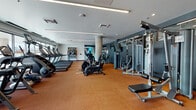 Located at 1420 Oak St in Lakewood, Colorado, this contemporary fitness center offers a comprehensive workout environment designed to support a wide range of exercise routines. The space is laid out across a single floor and features durable rubberized cork flooring throughout, providing both comfort and safety for high-impact activities.

The cardio zone is lined with multiple treadmills positioned along large floor-to-ceiling windows, allowing natural light to fill the area and offering views of the surrounding outdoor environment. Adjacent to this section, a dedicated cycling studio houses several stationary spin bikes alongside a large wall-mounted display screen and built-in cabinetry, creating a functional space for guided workout sessions.

The strength training area is well-appointed with a full array of cable machines, weight benches, free weights, and multi-functional rack systems, all set against a deep charcoal accent wall with integrated shelving for additional equipment storage. Mirrored walls enhance the sense of space and allow for form monitoring during workouts.

A functional training zone rounds out the facility, equipped with punching bags, kettlebells, medicine balls, battle ropes, and resistance tools neatly organized on wall-mounted racks. Architectural details such as linear recessed lighting, structural columns, and a clean neutral color palette give the space a polished, modern aesthetic throughout.
