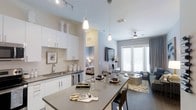 This contemporary single-floor residence showcases a thoughtfully designed open-concept layout that connects the kitchen, dining, and living areas in one cohesive space. The kitchen is outfitted with crisp white cabinetry, subway tile backsplash, a stainless steel sink, and integrated appliances, all complemented by a generous gray-topped island that doubles as a dining and prep space. Stylish pendant lights hang above the island, while track lighting illuminates the kitchen workspace. Soaring ceilings throughout amplify the sense of openness and airiness. The living area is anchored by a ceiling fan and large French-style doors that invite abundant natural light and provide a connection to the outdoors. Neutral tones in warm grays and whites create a clean, modern palette throughout. A flat-screen television is mounted on the wall, and a contemporary area rug with a subtle pattern grounds the living space. The bedroom is partially visible through an open passageway, hinting at the same refined aesthetic, with warm lighting and tasteful furnishings. Overall, the residence offers a bright, elegant, and functional environment suited for modern living.