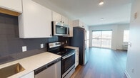 This single-floor residence showcases a contemporary design highlighted by warm hardwood-style flooring that runs consistently throughout the unit. The open-concept layout connects the kitchen directly to the living area, creating a cohesive and functional living space. The kitchen is well-appointed with stainless steel appliances, including an electric range, over-the-range microwave, dishwasher, and refrigerator. White cabinetry provides ample storage, while light-toned quartz-style countertops and a dark slate subway tile backsplash add a clean, modern aesthetic. The living area is bright and airy, benefiting from large windows and a sliding glass door that opens onto a private balcony, offering outdoor views of the surrounding area. Natural light flows freely through the space, enhancing its open feel. The bathroom features a full soaking tub with a shower, a white vanity with an undermount sink, a large mirror with overhead lighting, and a bold navy accent wall that adds visual interest. A dedicated utility closet houses an in-unit washer and includes wire shelving for additional storage. Overall, the unit presents a clean, modern aesthetic throughout.
