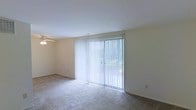 This single-floor residence offers a clean, functional layout with a comfortable and understated aesthetic. The main living area is a standout feature, bathed in natural light streaming through a large sliding glass door fitted with vertical blinds, which opens to an outdoor view and provides seamless indoor-outdoor connectivity. The space is finished with neutral, light-toned walls that contribute to an open and airy atmosphere. Underfoot, a combination of carpet and hard flooring adds subtle textural variety to the room. A ceiling fan is mounted overhead, ensuring efficient air circulation and year-round comfort. The overall design of the space is straightforward and practical, offering a blank canvas that is easy to personalize. The light color palette and generous natural light create a welcoming ambiance, making the living area feel bright and spacious. The single-floor layout provides convenience and accessibility throughout, and the sliding glass door enhances the sense of openness by visually extending the interior toward the outdoors.