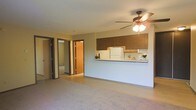 This single-floor space offers a functional and straightforward layout designed for comfortable everyday living. The living area is finished with wall-to-wall carpeting, creating a warm and quiet underfoot feel throughout the main space. A ceiling fan with an integrated light fixture is centrally positioned overhead, providing both air circulation and ambient lighting.

The kitchen is positioned along one wall and connects to the living area via a pass-through counter, which promotes an open feel while maintaining a defined cooking zone. Dark wood-toned upper cabinetry lines the kitchen wall, adding a traditional, grounded aesthetic. A white refrigerator is visible within the kitchen, and under-cabinet lighting provides task illumination for the workspace below.

Two doorways are visible from the main living area, suggesting access to additional rooms such as a bedroom and bathroom or storage areas. The doors feature warm wood-toned finishes consistent with the cabinetry, creating a cohesive interior palette. Neutral wall tones throughout keep the space feeling light and versatile. Overall, the layout is practical and efficiently organized for single-level living.