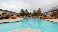This multi-building community is designed around a central outdoor courtyard that serves as its primary amenity hub. The buildings, finished in a neutral stucco exterior with dark rooflines, frame the outdoor space in a U-shaped configuration, creating an enclosed and cohesive environment. A prominent glass-fronted entrance structure anchors the central building, adding an architectural focal point to the courtyard.

The outdoor area is well-equipped for recreation and relaxation. A generously sized swimming pool with a decorative tiled motif and a natural rock water feature serves as the centerpiece, complemented by a separate circular hot tub nearby. Blue lounge chairs line the pool deck, offering ample space for sunbathing. An open lawn area provides green space at the heart of the courtyard, bordered by a curved concrete walkway. A raised fire pit adds a communal gathering element to the hardscaped surroundings.

Outdoor dining tables and chairs are positioned along the patio, suitable for casual gatherings. Mature pine trees, ornamental shrubs, and mulched planting beds line the perimeter, providing privacy and greenery. A gated wrought-iron entrance with a keypad offers controlled access to the community, enhancing security and privacy throughout.