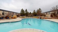 This multi-building community is designed around a central outdoor courtyard that serves as its primary amenity hub. The buildings, finished in a neutral stucco exterior with dark rooflines, frame the outdoor space in a U-shaped configuration, creating an enclosed and cohesive environment. A prominent glass-fronted entrance structure anchors the central building, adding an architectural focal point to the courtyard.

The outdoor area is well-equipped for recreation and relaxation. A generously sized swimming pool with a decorative tiled motif and a natural rock water feature serves as the centerpiece, complemented by a separate circular hot tub nearby. Blue lounge chairs line the pool deck, offering ample space for sunbathing. An open lawn area provides green space at the heart of the courtyard, bordered by a curved concrete walkway. A raised fire pit adds a communal gathering element to the hardscaped surroundings.

Outdoor dining tables and chairs are positioned along the patio, suitable for casual gatherings. Mature pine trees, ornamental shrubs, and mulched planting beds line the perimeter, providing privacy and greenery. A gated wrought-iron entrance with a keypad offers controlled access to the community, enhancing security and privacy throughout.