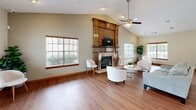 Located at 1200 Lanier Mill Circle in Oakwood, Georgia, this single-story community clubhouse offers a well-appointed and inviting gathering space. The open-concept layout is anchored by a spacious lounge area featuring warm hardwood-style flooring, vaulted ceilings with a ceiling fan, and a striking wood-mantled fireplace with tile surround serving as the room's focal point. Multiple windows fitted with blinds allow abundant natural light to fill the space, creating a bright and airy atmosphere throughout. The lounge comfortably accommodates multiple seating arrangements, making it ideal for both casual gatherings and community events. French glass doors at the entry provide an elegant welcome and connect the interior to the exterior. Adjacent to the lounge is a fully functional kitchen, equipped with light wood cabinetry, stainless steel appliances including a refrigerator and microwave, tile flooring, and generous counter space. A peninsula-style breakfast bar with seating for multiple guests bridges the kitchen and the main lounge area, encouraging social interaction. Crown molding and warm wood accents throughout the space lend a cohesive, polished finish. The overall ambiance is comfortable, functional, and well-suited for community use.