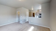 This single-floor residence offers a clean, functional layout designed for comfortable everyday living. The main living area features wall-to-wall carpet in a neutral tone, contributing to a warm and inviting atmosphere. Soft beige walls and flush-mount ceiling lighting create a bright, evenly lit environment throughout the space.

The kitchen is positioned adjacent to the living area and is defined by a raised peninsula counter that provides a natural division between the two spaces while maintaining an open feel. Light wood cabinetry offers ample storage, and the kitchen is equipped with stainless steel appliances, including a microwave and a range, set against a crisp white backsplash. A pass-through opening connects the kitchen to a secondary storage or pantry area beyond.

Two interior doors are visible from the living space — one featuring a paneled design that leads to an additional room or hallway, and another that opens to a closet or utility area. The thoughtful floor plan maximizes the use of available space while keeping circulation paths clear and accessible. Overall, the residence presents a tidy, neutral aesthetic that offers a versatile canvas for personalization.