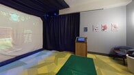 Located at 1600 Harbor Blvd in Weehawken, NJ, this single-floor sports simulation space offers an engaging and tech-forward recreational experience. The main bay is defined by a large projection screen framed by floor-to-ceiling black drape curtains, which serve both a functional and atmospheric purpose by focusing attention on the immersive simulator display. A ceiling-mounted projector delivers sharp visuals for a variety of sports simulations — including lacrosse and golf — as evidenced by the high score leaderboard and sport-selection interface visible on screen. A central kiosk with a touchscreen interface allows users to navigate and select their sport of choice, clearly numbered and well-positioned for easy access. The flooring features a vibrant geometric-patterned carpet in shades of green and yellow, with a dedicated synthetic turf mat serving as the active play area. Accent wall sconces cast a warm glow against the gray walls, complemented by artwork that adds a light, sporty character to the space. A small seating arrangement with modern bucket chairs and a side table provides a comfortable waiting or spectating area near the entrance. The space is thoughtfully designed to balance active play with practical amenities.