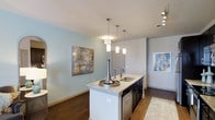 This single-floor residence in Texas offers a thoughtfully designed open-concept layout that seamlessly connects the kitchen and living areas, creating a welcoming and functional environment. The kitchen serves as the heart of the space, anchored by a sleek center island topped with light granite countertops and a stainless steel sink. Pendant lighting in varying cylindrical styles hangs elegantly above the island, providing both task lighting and a refined aesthetic focal point. Dark cabinetry beneath the island contrasts beautifully with the light countertop surfaces, while stainless steel appliances, including a dishwasher, add a contemporary edge. Track lighting along the ceiling further enhances the kitchen's illumination. The flooring throughout features rich, dark hardwood, grounding the space with warmth and sophistication. Soft, light-toned walls in a cool blue-gray hue lend the interior a calm, airy ambiance. Decorative artwork adds character to the walls, complementing the modern aesthetic. The open sightlines throughout the main living area create a generous sense of space, making this residence feel both comfortable and stylish. Overall, the space strikes a balance between modern design and everyday practicality.