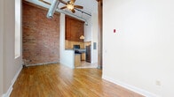 Located at 79 Warren St in Glens Falls, NY, this efficiently designed single-floor unit blends industrial character with functional living. The open-concept kitchen and living area is anchored by soaring ceilings that amplify the sense of space, with exposed ductwork, a ceiling fan, and original wooden structural beams lending an authentic loft-style aesthetic. Exposed brick walls add warmth and texture throughout, creating a striking visual contrast against the clean white walls and warm hardwood flooring.

The kitchen features light maple cabinetry, granite-style countertops, and a full suite of stainless steel appliances including a refrigerator, electric range, built-in microwave, and dishwasher. Tile flooring grounds the kitchen area, while the open layout connects seamlessly to the living space. Large multi-pane windows along the living area wall invite an abundance of natural light and offer views of the surrounding streetscape.

The bathroom is clean and straightforward, appointed with a pedestal sink, a tub-shower combination, tile flooring, and a medicine cabinet mirror. Throughout the unit, the high ceilings, industrial details, and natural light combine to create an urban yet inviting atmosphere well-suited to city living in downtown Glens Falls.