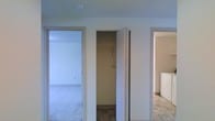 This single-floor space is characterized by a practical and straightforward layout, centered around a hallway that connects its key areas. The hallway serves as a functional hub, featuring a dedicated closet equipped with a hanging rod and shelving, providing convenient built-in storage. The closet is finished with tile flooring, adding a durable surface underfoot.

To one side of the hallway, a doorway opens into a carpeted room bathed in soft, diffused natural light, suggesting a comfortable living or sleeping area. The neutral white walls and clean lines of the space contribute to a bright and open atmosphere. On the opposite side, a passage leads to what appears to be a kitchen or utility area, identifiable by its mixed tile flooring and a glimpse of white cabinetry, hinting at a functional and organized space.

Throughout, the interiors are finished in a consistent neutral palette of whites and soft grays, lending a cohesive and understated aesthetic. The layout reflects a straightforward, efficient design, making thoughtful use of the available space across its single level.