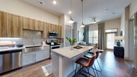This well-appointed single-floor residence showcases a thoughtfully designed open-concept layout that seamlessly connects the kitchen, dining, and living areas. The gourmet kitchen is a standout feature, equipped with stainless steel appliances, a farmhouse-style sink, a shimmering tile backsplash, and a generous granite-topped island with bar seating illuminated by stylish pendant lighting. Warm wood-tone upper cabinetry contrasts elegantly with the light lower cabinets, while rich hardwood-style flooring flows throughout the main living areas. The living room benefits from soaring ceilings and floor-to-ceiling windows that flood the space with natural light and provide views to an outdoor balcony. Two well-sized bedrooms offer carpeted comfort, ample natural light, and generous closet space. The residences bathrooms are finished with granite countertops — one featuring a vibrant blue vanity with a tub-shower combination, and another with clean grey cabinetry and a walk-in shower. Throughout the home, neutral tones create a versatile backdrop, while high ceilings enhance the sense of openness. The cohesive design balances modern finishes with warm, inviting details throughout every room.