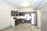 This single-floor space showcases a thoughtfully designed interior with a cohesive modern aesthetic throughout. The kitchen serves as a standout feature, outfitted with dark wood-grain cabinetry, white quartz countertops, and a full suite of stainless steel appliances including a range, over-the-range microwave, dishwasher, and refrigerator. A deep stainless steel farmhouse sink and classic white subway tile backsplash add both practicality and style to the cooking space. Polished terrazzo flooring extends throughout the unit, lending a durable yet refined character to each room. The main living area benefits from natural light streaming through double windows dressed with horizontal blinds, creating a bright and welcoming atmosphere. A sliding door adds a space-conscious design element while maintaining an open flow between rooms. The bathroom is well-appointed with a soaking tub set against large-format gray tile walls in a linear pattern, complemented by chrome fixtures and a curtain rod. The overall design palette — neutral whites, warm grays, and dark cabinetry accents — creates a clean, contemporary environment that is both functional and visually cohesive.