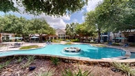 Located at 1000 Farrah Ln in Stafford, Texas, this outdoor amenity space offers a richly landscaped retreat designed for relaxation and recreation. At its centerpiece sits a generously sized, freeform swimming pool with crystal-clear water, flanked by a built-in spa and an ornamental tiled fountain that adds a decorative focal point to the grounds. The pool deck is finished in aggregate concrete and furnished with lounge chairs, umbrella-shaded tables, and seating areas ideal for outdoor gatherings.

Mature crape myrtles and large shade trees are planted throughout the space, creating a lush, park-like canopy that provides natural shade and privacy. Stone-capped retaining walls and curved walkways guide visitors through the landscaped grounds, with native shrubs and planted beds adding layers of greenery. A wood-framed pergola offers additional shaded seating and serves as a gathering space within the complex.

The surrounding residential buildings feature a combination of brick and blue-toned siding with private balconies overlooking the courtyard. Wrought-iron fencing encloses the amenity area, maintaining a secure and well-defined perimeter. The overall ambiance is that of a thoughtfully designed outdoor community space that balances functional amenities with attractive, resort-inspired landscaping.