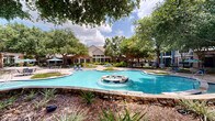 Located at 1000 Farrah Ln in Stafford, Texas, this outdoor amenity space offers a richly landscaped retreat designed for relaxation and recreation. At its centerpiece sits a generously sized, freeform swimming pool with crystal-clear water, flanked by a built-in spa and an ornamental tiled fountain that adds a decorative focal point to the grounds. The pool deck is finished in aggregate concrete and furnished with lounge chairs, umbrella-shaded tables, and seating areas ideal for outdoor gatherings.

Mature crape myrtles and large shade trees are planted throughout the space, creating a lush, park-like canopy that provides natural shade and privacy. Stone-capped retaining walls and curved walkways guide visitors through the landscaped grounds, with native shrubs and planted beds adding layers of greenery. A wood-framed pergola offers additional shaded seating and serves as a gathering space within the complex.

The surrounding residential buildings feature a combination of brick and blue-toned siding with private balconies overlooking the courtyard. Wrought-iron fencing encloses the amenity area, maintaining a secure and well-defined perimeter. The overall ambiance is that of a thoughtfully designed outdoor community space that balances functional amenities with attractive, resort-inspired landscaping.