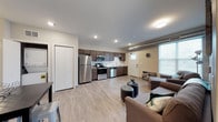 This single-floor space showcases a thoughtfully designed open-concept layout that seamlessly integrates the kitchen and living area into one cohesive, functional environment. The light hardwood-style flooring flows throughout, creating a sense of continuity and warmth across the entire space.

The kitchen is well-appointed with dark espresso cabinetry, stainless steel appliances including a full-sized refrigerator, a gas range, a built-in microwave, and a dishwasher. A clean white countertop provides ample workspace, and the layout is designed for both efficiency and style. A door near the kitchen offers direct access to the exterior, adding convenience.

The living area is generously proportioned, offering comfortable room for seating arrangements and everyday living. Recessed lighting throughout the space ensures consistent, ambient illumination, complemented by natural light streaming in through a large window. The neutral white walls and ceiling enhance the brightness of the interior and provide a clean backdrop.

Exposed ductwork along the upper wall adds an industrial accent that contrasts nicely with the contemporary finishes. A closet near the entrance provides practical storage. Overall, the space strikes a balance between modern aesthetics and functional design, making it an inviting and practical environment.