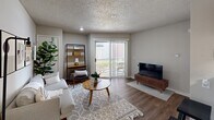 This 1-bedroom, 1-bathroom unit offers a thoughtfully organized single-floor layout comprising a living room, bedroom, kitchen, bathroom, and hallway. The living room serves as the heart of the space, featuring wood-look flooring, a textured ceiling, and a sliding glass door that opens to an outdoor balcony, filling the room with natural light and providing outdoor views. The galley-style kitchen is efficiently designed with warm oak cabinetry, white countertops, black appliances including a gas range and refrigerator, and a stainless steel sink — maximizing functionality within a compact footprint. The bedroom is a comfortable, well-proportioned retreat equipped with a ceiling fan, wood-look flooring, a textured ceiling, and a window dressed with blinds and dark curtains. The bathroom features a wood-toned vanity with a wide mirror, a three-bulb vanity light fixture, an overhead medicine cabinet, and a shower-tub combination with a curtain. Neutral gray tones throughout the walls create a clean, cohesive aesthetic that ties each room together, while consistent wood-look flooring adds warmth and continuity across the space.