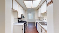 Located at 18418 Dearborn St in the Northridge neighborhood of Los Angeles, this single-story residence offers a well-organized layout designed for comfortable everyday living. The galley-style kitchen is efficiently arranged with white cabinetry lining both walls, light countertops, a stainless steel sink, a gas range with built-in microwave, and a black dishwasher. A skylight panel overhead fills the kitchen with bright, diffused natural light, while rich hardwood-style flooring runs throughout. The living area is a standout feature, boasting vaulted ceilings with exposed beams, a fireplace, and a high clerestory window that floods the room with abundant sunlight. The bedrooms are carpeted and bright, each with windows fitted with vertical blinds and built-in closets providing ample storage. Two bathrooms serve the home: one features a shower-tub combination with sliding glass doors, a large vanity mirror, and a single-sink vanity with white cabinetry; the second offers a similarly appointed vanity with generous counter space. Consistent white walls and neutral tones throughout create a clean, airy atmosphere that is both functional and inviting.