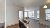 This single-floor space showcases an open-concept layout that seamlessly connects the kitchen and living areas. The kitchen island serves as a focal point, featuring a granite or stone-composite countertop, a double-basin stainless steel sink with a curved bronze-finish faucet, and dark espresso-toned cabinetry underneath. A single pendant light hangs above the island, adding a warm, focused glow to the workspace.

The living area extends naturally from the kitchen, finished with neutral-toned carpeting that contrasts with the hardwood-style flooring in the kitchen zone. Multiple windows fitted with horizontal blinds line the far wall, allowing generous natural light to fill the space and create a bright, airy atmosphere. The clean white walls and smooth ceilings contribute to the overall sense of openness.

A doorway visible from the kitchen leads to an adjacent room, suggesting additional private spaces beyond the main open-plan area. The overall design balances functional kitchen elements with a comfortable, light-filled living space, making for a practical and cohesive interior.