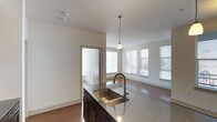 This single-floor space showcases an open-concept layout that seamlessly connects the kitchen and living areas. The kitchen island serves as a focal point, featuring a granite or stone-composite countertop, a double-basin stainless steel sink with a curved bronze-finish faucet, and dark espresso-toned cabinetry underneath. A single pendant light hangs above the island, adding a warm, focused glow to the workspace.

The living area extends naturally from the kitchen, finished with neutral-toned carpeting that contrasts with the hardwood-style flooring in the kitchen zone. Multiple windows fitted with horizontal blinds line the far wall, allowing generous natural light to fill the space and create a bright, airy atmosphere. The clean white walls and smooth ceilings contribute to the overall sense of openness.

A doorway visible from the kitchen leads to an adjacent room, suggesting additional private spaces beyond the main open-plan area. The overall design balances functional kitchen elements with a comfortable, light-filled living space, making for a practical and cohesive interior.
