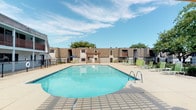 Located at 3201 E Park Row Dr in Arlington, Texas, this outdoor amenity space is designed to offer residents a well-equipped and inviting environment for relaxation and outdoor entertaining. The centerpiece of the space is a generously sized swimming pool with clear blue water, framed by a spacious concrete pool deck furnished with green lounge chairs, providing ample room for sunbathing under the Texas sky. A black metal security fence encloses the pool area, ensuring a safe and defined perimeter. Adjacent to the pool, a substantial wood pergola shelters an outdoor kitchen station featuring a stainless steel built-in grill set into a stone-clad counter with a granite surface — a standout feature ideal for outdoor gatherings. Casual patio seating with a table, chairs, and a blue market umbrella adds a comfortable social area nearby. Mature trees provide natural shade and greenery throughout the courtyard, while well-maintained landscaped beds and grassy areas contribute to the overall appeal. The surrounding buildings exhibit a distinctly mid-century architectural character, with brick and shake-style siding, angled rooflines, and arched accent details. The space benefits from abundant natural light and open sky views, creating a bright and welcoming outdoor retreat.