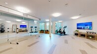 Located at 1001 N Coast Highway in Oceanside, California, this contemporary fitness studio offers a well-appointed, single-floor exercise space designed to accommodate a variety of workout disciplines. The interior is defined by a clean, minimalist aesthetic, with crisp white walls, smooth white ceilings, and light wood-toned flooring that lends the studio an open, airy feel.

A full wall of floor-to-ceiling mirrors lines one side of the studio, visually expanding the space and providing essential feedback during workouts. Freestanding ballet barres positioned in front of the mirrors make the space well-suited for barre, dance, and stretching classes. On the cardio side, Peloton spin bikes are stationed on protective mats, each equipped with interactive screens for guided cycling sessions.

Strength and functional training are supported by a rack of dumbbells, kettlebells, stability balls, BOSU trainers, and step platforms, offering versatile workout options. A wall-mounted flat-screen TV serves as an additional media resource for fitness programming. Large floor-to-ceiling windows along one wall flood the studio with natural light and provide views of surrounding greenery, creating a bright and energizing atmosphere. Recessed and flush-mount ceiling lights ensure consistent illumination throughout. The space also includes a full-length standing mirror and storage solutions for equipment organization.