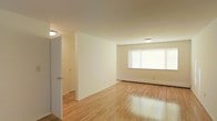 This two-floor space presents a clean, functional layout characterized by light-toned hardwood flooring that runs throughout the main living area, lending warmth and continuity to the interior. The walls are finished in a neutral off-white palette, creating a bright and airy atmosphere that complements the natural light entering through a large window along one wall. The window is fitted with white blinds, allowing for adjustable light control while maintaining a streamlined appearance.

The main living area is open and generously proportioned, offering flexibility in how the space can be arranged and utilized. A partial wall divides the living space from an adjacent hallway or secondary area, adding a degree of structural definition without fully enclosing the layout. This hallway leads to additional rooms, suggesting a practical flow between spaces.

Ceiling-mounted flush light fixtures provide overhead illumination, supplementing the natural light available. A baseboard heating unit runs along the lower wall beneath the window, indicating a reliable heating system integrated into the architecture. The overall ambiance is understated and neutral, providing a versatile foundation suitable for a variety of uses across its two floors.