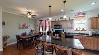This single-floor home offers a thoughtfully designed layout that balances everyday functionality with comfortable living. The kitchen is a standout feature, showcasing warm medium-tone wood cabinetry, dark granite countertops, and black appliances including a refrigerator, range, and built-in microwave. A large central island with seating provides additional prep space and casual dining, while pendant lighting adds character overhead. The adjacent dining area accommodates a full dining table set beneath a ceiling fan, with natural light filtering through windows dressed in rich red curtains. Crown molding adds a refined architectural detail throughout the main living areas. The living room is generously proportioned, featuring rich hardwood flooring, a large sectional sofa, and a bank of windows that floods the space with natural light. Two bathrooms serve the home — one full bath with a tub and shower combination, a single-sink vanity with wood cabinetry, and tile flooring, and a second half bath with matching tile and a compact vanity. The bedrooms are carpeted and well-lit, with multiple windows offering natural brightness. One bedroom features neutral tones and ample space, while another serves as a versatile secondary room. Overall, the home presents a warm, lived-in atmosphere with consistent finishes across every room.