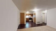 This single-floor unit presents an open-concept layout that connects the living area to the kitchen, creating a cohesive and functional flow throughout the space. The living area features neutral carpet flooring, providing a comfortable underfoot feel, while the kitchen and adjoining hallway areas showcase dark hardwood-style flooring that adds contrast and visual interest.

The kitchen is equipped with warm, medium-toned wooden cabinetry arranged in an L-shaped configuration, offering ample storage. White appliances, including a range and oven, are integrated neatly beneath the upper cabinets. A flush-mounted ceiling light illuminates the kitchen area, complementing the under-cabinet lighting above the range.

Multiple interior doors are visible throughout the space, suggesting access to additional rooms or storage areas. A floor-mounted air vent indicates a forced-air climate system. The walls are finished in a clean, neutral white throughout, contributing to a bright and airy atmosphere. Overall, the space maintains a practical, well-organized design with a simple and understated aesthetic.
