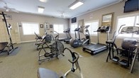 This fitness center offers a comprehensive workout environment housed within a single-floor layout. The room is finished with neutral-toned walls and low-pile carpeted flooring, creating a clean and functional atmosphere conducive to exercise. Recessed panel lighting and a ceiling fan ensure the space remains well-lit and comfortably ventilated throughout the day. Multiple windows along the walls allow generous natural light to fill the room, enhancing the overall ambiance. The facility is outfitted with a diverse selection of cardio equipment, including treadmills, an elliptical trainer, a stationary upright bike, an air resistance bike, and a stair-stepper, catering to a wide range of fitness preferences and workout routines. A wall-mounted flat-screen television is positioned for easy viewing during exercise sessions. A large mirror along one wall adds a sense of depth and openness to the room while serving a practical purpose for form monitoring. Framed artwork and motivational pieces decorate the walls, adding a welcoming touch to the utilitarian space. Informational and safety signage is also displayed, reflecting a commitment to user safety and guidance. Overall, this fitness center provides a well-rounded and accessible environment for cardiovascular training and general exercise.