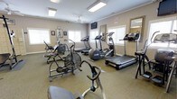 This fitness center offers a comprehensive workout environment housed within a single-floor layout. The room is finished with neutral-toned walls and low-pile carpeted flooring, creating a clean and functional atmosphere conducive to exercise. Recessed panel lighting and a ceiling fan ensure the space remains well-lit and comfortably ventilated throughout the day. Multiple windows along the walls allow generous natural light to fill the room, enhancing the overall ambiance. The facility is outfitted with a diverse selection of cardio equipment, including treadmills, an elliptical trainer, a stationary upright bike, an air resistance bike, and a stair-stepper, catering to a wide range of fitness preferences and workout routines. A wall-mounted flat-screen television is positioned for easy viewing during exercise sessions. A large mirror along one wall adds a sense of depth and openness to the room while serving a practical purpose for form monitoring. Framed artwork and motivational pieces decorate the walls, adding a welcoming touch to the utilitarian space. Informational and safety signage is also displayed, reflecting a commitment to user safety and guidance. Overall, this fitness center provides a well-rounded and accessible environment for cardiovascular training and general exercise.