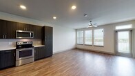 Located at 1700 W Frankford Rd in Carrollton, Texas, this single-floor unit offers a thoughtfully designed open-concept layout that seamlessly connects the kitchen and living area. Warm wood-tone flooring runs throughout the space, complementing the clean, modern aesthetic.

The kitchen is a standout feature, outfitted with dark espresso cabinetry reaching toward the ceiling for ample storage, crisp white countertops, and a full suite of stainless steel appliances including a refrigerator, range, microwave, and dishwasher. A subway tile backsplash adds a polished finishing touch.

The spacious living area is bathed in natural light from a bank of large windows fitted with blinds, offering outdoor views and a bright, airy atmosphere. A ceiling fan provides additional comfort, and recessed lighting illuminates the space throughout.

The bathroom continues the modern design language with dark wood cabinetry, a wide white countertop vanity, a large mirror, and a full soaking tub with clean white tile surround. Wood-look flooring extends into this room as well, tying the unit together cohesively.

Overall, this unit presents a contemporary and functional living environment with consistent, high-quality finishes and a well-utilized single-floor floor plan.