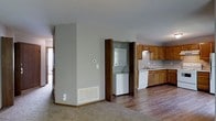 This single-floor residence offers a practical and well-organized layout designed for comfortable everyday living. The open-concept design connects the living area to the kitchen, creating a seamless flow throughout the main living space. The kitchen is equipped with warm honey-toned wood cabinetry, a double sink, and white appliances, including a dishwasher, all set atop durable hardwood-style flooring. Under-cabinet space and a ventilation hood above the range add to the kitchen's functionality. A standout convenience of this home is the dedicated in-unit laundry closet, which houses a stacked washer and dryer unit tucked neatly behind bifold doors — a practical feature that adds significant value. The living area transitions to plush carpeting, offering a cozy contrast to the hard flooring in the kitchen zone. Neutral gray walls throughout lend the space a clean, understated aesthetic that is easy to personalize. A floor vent provides in-floor heating, and a thermostat is conveniently positioned on the wall. Natural light filters in from an adjacent room visible in the background, contributing to an airy and welcoming atmosphere throughout this efficiently designed single-level home.