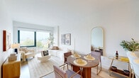 This thoughtfully designed 2-bedroom, 1-bathroom residence is laid out across a single floor, offering a cohesive and efficient use of space. The open-plan kitchen and living area forms the heart of the home, featuring warm natural wood cabinetry, light-toned open shelving, white quartz countertops, and a full suite of stainless steel appliances including a French door refrigerator, electric range, and built-in microwave. A peninsula with seating creates a casual dining option, seamlessly connecting the kitchen to the living area. The living room is bright and airy, anchored by a large picture window framing expansive city and rooftop views, complemented by light hardwood flooring that extends throughout. The primary bedroom is a serene retreat with large windows offering the same sweeping views, plush carpeting, and ample natural light. The second bedroom maintains the same understated, neutral aesthetic. A well-appointed hallway with additional closet storage connects the rooms with ease. The bathroom is finished with large-format tile flooring, a sleek navy floating vanity, an illuminated mirror cabinet, and clean, modern fixtures. Overall, the space presents a calm, contemporary ambiance with a consistent palette of whites, warm woods, and soft neutrals.