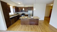 This single-floor space centers around a well-appointed kitchen designed for both functionality and style. The kitchen showcases rich dark wood cabinetry that extends to the upper walls, providing generous storage throughout. Stainless steel appliances, including a refrigerator, electric range, and built-in microwave, complement the cabinetry with a sleek, modern aesthetic.

A central kitchen island serves as a focal point, featuring a granite countertop with subtle warm tones and seating for multiple guests, making it ideal for casual dining or entertaining. Pendant lights hang above the island, adding targeted illumination and a design accent that ties the space together.

The countertops along the perimeter mirror the granite of the island, maintaining a cohesive look throughout the kitchen. Warm-toned hardwood flooring extends from the kitchen into the adjacent living area, creating a seamless visual flow between spaces. A sink positioned near a window allows for natural light in the workspace. The overall ambiance is warm and inviting, balancing practical kitchen functionality with contemporary design elements.