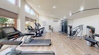 This fitness center in Irvine, CA offers a well-equipped exercise space designed to support a comprehensive workout routine. The room features light hardwood-style flooring throughout, crisp white walls, and recessed lighting that creates a bright and clean atmosphere. High clerestory windows run along two sides of the room, flooding the space with abundant natural light and offering views of the surrounding outdoor areas, including a pool and landscaped grounds visible through large glass doors and windows. The cardio section is well-stocked with a row of treadmills and elliptical machines from Matrix, providing ample options for aerobic exercise. The strength training area includes a functional cable cross trainer, a multi-use weight machine, a flat bench, and additional selectorized equipment for targeted muscle group training. A water fountain and wall-mounted air conditioning unit ensure comfort during workouts. Fitness center rules are posted prominently on the wall, maintaining an organized and community-friendly environment. A direct door leads to the outdoor pool area, seamlessly connecting indoor fitness with outdoor recreation. The overall ambiance is modern, airy, and purpose-driven, making it an inviting space for residents to maintain an active lifestyle.