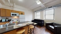 Located at 5 Lyon St NW in downtown Grand Rapids, Michigan, this single-floor unit showcases a blend of urban industrial design and functional living. The open-concept layout seamlessly connects the kitchen and living area, creating a cohesive and practical space. The kitchen features warm wood cabinetry, a full suite of white appliances including a refrigerator, electric range, and microwave, all centered around a peninsula with a dark countertop and built-in seating for two — ideal for casual dining. Hardwood flooring runs throughout the main living area, adding warmth and contrast to the bright white walls. The living room is brightened by two large windows fitted with blinds, allowing ample natural light to fill the space. Exposed industrial-style ductwork runs along the ceiling, lending the unit a distinctive loft-like character. A private bedroom offers carpeted flooring and a quieter retreat, also featuring exposed ductwork overhead. The bathroom is cleanly appointed with tile flooring, a single-sink vanity with wood cabinetry, and a full shower-tub combination. The overall ambiance is modern and utilitarian, with urban design elements that reflect the building's character in the heart of Grand Rapids.