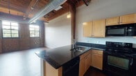 Located at 1119 Mississippi Ave in St. Louis, MO, this industrial-style loft unit showcases a compelling blend of historic character and modern finishes throughout its single-floor layout. The open-concept living area is defined by soaring timber beam ceilings with exposed ductwork and track lighting, lending a raw, warehouse aesthetic that is both functional and visually striking. Original exposed brick walls line the main living space, accompanied by large multi-pane windows that flood the interior with abundant natural light and offer views of the surrounding neighborhood.

The kitchen is well-appointed with light maple cabinetry, dark granite countertops, and a full suite of black appliances including a range, microwave, dishwasher, and refrigerator. A peninsula provides additional counter space and serves as a natural division between the kitchen and the expansive living area, which features warm hardwood-style flooring throughout.

The bedroom continues the loft's industrial character with exposed brick accents, a ceiling fan, and plush carpet for added comfort. An en-suite bathroom offers a vanity with dark granite countertops, maple cabinetry, a large mirror, a soaking tub, and a separate closet with convenient access. The overall ambiance is one of urban character paired with thoughtful, livable design.