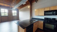 Located at 1119 Mississippi Ave in St. Louis, MO, this industrial-style loft unit showcases a compelling blend of historic character and modern finishes throughout its single-floor layout. The open-concept living area is defined by soaring timber beam ceilings with exposed ductwork and track lighting, lending a raw, warehouse aesthetic that is both functional and visually striking. Original exposed brick walls line the main living space, accompanied by large multi-pane windows that flood the interior with abundant natural light and offer views of the surrounding neighborhood.

The kitchen is well-appointed with light maple cabinetry, dark granite countertops, and a full suite of black appliances including a range, microwave, dishwasher, and refrigerator. A peninsula provides additional counter space and serves as a natural division between the kitchen and the expansive living area, which features warm hardwood-style flooring throughout.

The bedroom continues the loft's industrial character with exposed brick accents, a ceiling fan, and plush carpet for added comfort. An en-suite bathroom offers a vanity with dark granite countertops, maple cabinetry, a large mirror, a soaking tub, and a separate closet with convenient access. The overall ambiance is one of urban character paired with thoughtful, livable design.