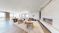 Located at 1 Gibson Way in Revere, MA, this generously sized amenity floor spans an impressive footprint and showcases a cohesive modern design throughout. The living room is a standout centerpiece, featuring wide-plank hardwood floors, a sleek linear fireplace framed in stone, floor-to-ceiling windows that flood the space with natural light, and views of the surrounding area. Contemporary furnishings and a neutral palette create a welcoming, sophisticated ambiance.

The reception and lounge area is anchored by a curved white island with bar seating, complemented by warm wood-slat accent walls, potted greenery, and elegant recessed lighting. A dramatic open stairwell with curved white balustrades, a coffered ceiling with ambient LED uplighting, and lush indoor plantings serves as an architectural focal point connecting the floor's various zones.

The fitness center is outfitted with a full row of commercial-grade treadmills and cardio equipment, floor-to-ceiling glass partitions, and rubber tile flooring. An adjacent studio features sprung hardwood floors, full-length mirrors, moss wall panels, and a mounted display screen — ideal for yoga or group fitness. A private wellness room offers a massage chair, artistic wallcovering, and a serene retreat atmosphere. Wide hallways, clean white walls with curated artwork, and consistent hardwood flooring unify the entire floor.