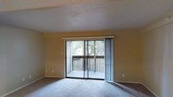 This single-floor space features a carpeted living area finished in neutral tones, creating a clean and understated atmosphere. The room is defined by large sliding glass doors that span a significant portion of one wall, allowing natural light to filter in and providing direct access to a private balcony. The balcony is enclosed by a slatted railing, beyond which mature trees are visible, offering a sense of natural surroundings and privacy. The interior walls are painted in a warm beige tone, complementing the light-colored carpet underfoot. The ceiling features a textured finish and is framed by a subtle architectural soffit detail along one side of the room, adding a modest structural accent. Vertical blinds accompany the sliding doors, allowing for adjustable light control and privacy. Electrical outlets are positioned at baseboard level around the room, and the overall layout suggests a straightforward, functional design. The combination of natural light, outdoor access, and neutral finishes gives the space a calm, livable character suited for everyday use.