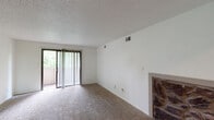 This single-story unit offers a functional and comfortable layout spread across several well-defined areas. The living room serves as the central gathering space, featuring neutral-toned wall-to-wall carpeting, a textured ceiling, and a notable stone fireplace that adds a rustic focal point to the room. A sliding glass door fitted with vertical blinds opens to an outdoor balcony, allowing natural light to filter in and providing views of surrounding greenery. The entry foyer transitions smoothly from tile flooring into the carpeted living spaces, offering a clear delineation between zones.

The kitchen is equipped with dark mahogany-toned cabinetry offering generous upper and lower storage, light-toned countertops, and a full suite of black appliances including a gas range, over-the-range microwave, dishwasher, and refrigerator. A double sink and tile flooring complete the practical workspace.

The bedrooms are carpeted and finished with textured ceilings. At least one bedroom features a ceiling fan with integrated lighting, adding to the comfort of the space. Closet access is visible in the bedroom hallway, along with proximity to a bathroom. Throughout, the unit maintains a clean, neutral palette with white walls, creating a bright and adaptable interior environment.