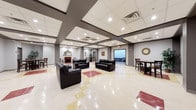 This single-floor space functions as a generously sized common gathering area, designed to accommodate both relaxed lounging and casual social interaction. The interior is anchored by a striking coffered ceiling with dark-framed beams that create a sense of architectural depth, complemented by recessed lighting and flush panel tiles that illuminate the space evenly throughout.

The flooring is a polished composite tile in cream, beige, and deep red tones, lending a clean and refined aesthetic to the open layout. A seating area features multiple black leather sofas and chairs arranged to encourage conversation, positioned near a decorative fireplace with a classic mantel and mirror—adding a warm, traditional focal point to the room.

Toward one end of the space, a windowed opening provides a glimpse of an outdoor view, drawing in natural light. To the side, a dining or café-style area includes dark wood high-top tables and matching chairs, ideal for informal gatherings. A decorative wall clock adds a functional yet stylish detail to the space. The overall ambiance is open, well-lit, and purposefully designed for communal use.