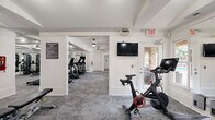 This fitness center offers a comprehensive workout facility spread across a single floor, thoughtfully organized into distinct activity zones. The space features a clean, modern aesthetic characterized by crisp white walls, coffered ceilings with decorative beams, and stylish pendant and flush-mount lighting that keeps the environment bright and energizing.

The cardio zone is equipped with multiple treadmills and a Peloton spin bike, accompanied by wall-mounted flat-screen TVs for entertainment during workouts. Large mirrors line the walls throughout, amplifying the sense of space and allowing users to monitor their form. Full-length glass doors provide views to the exterior and invite natural light deep into the facility.

The strength training area is generously stocked with a multi-station cable machine, a rack of kettlebells and dumbbells, stability balls, and a weight bench, offering a well-rounded selection of free weights and functional training tools. The patterned gray carpet flooring provides both comfort and traction underfoot.

A dedicated spin and stretching zone near the entrance features additional cardio equipment and a mounted display screen. Fitness center rules are clearly posted, and safety features including emergency exit signage are prominently placed. The open layout and interconnected rooms create a seamless, functional flow throughout the entire facility.