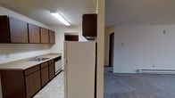 This single-floor residence offers a practical and straightforward layout designed for comfortable everyday living. The kitchen is equipped with dark wood cabinetry that provides ample storage space, a double-basin sink, a dishwasher, and a full-size refrigerator. The countertops are light-colored and complement the tile flooring underfoot, which is easy to maintain and clean. Overhead fluorescent lighting brightens the kitchen workspace effectively. A structural column with an overhead cabinet serves as a natural divider between the kitchen and the adjacent living area, creating a sense of separation while maintaining an open feel. The living area features wall-to-wall carpeting in a neutral gray tone, lending a softer, more comfortable atmosphere to the space. A hallway visible from the main area leads to additional rooms, suggesting a well-organized floor plan. The textured ceilings and neutral wall colors throughout contribute to a clean, understated aesthetic. Overall, the space is practical and move-in ready, offering a cohesive flow between its kitchen and living areas.